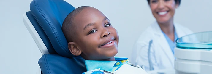 Young boy smiling comfortably during his first dental visit, with pediatric dentist in background. Young boy smiling comfortably during his first dental visit, with pediatric dentist in background.