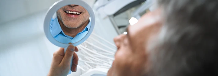 A male patient with a stubbled jaw admires his new dental implants smiling back at him in a hand held mirror A male patient with a stubbled jaw admires his new dental implants smiling back at him in a hand held mirror