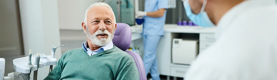 An older bearded gentelman in a collared shirt with a seafoam green pullover sits in the exam chair while a dental assistant preps in the background out of focus