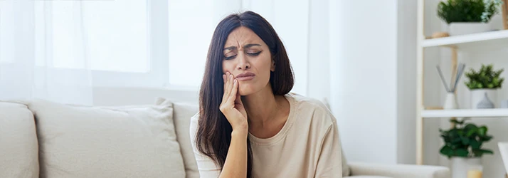 A woman with long dark brown hair over her shoulder firmly presses her fingers to her clenched jaw, brow furrowed in pain A woman with long dark brown hair over her shoulder firmly presses her fingers to her clenched jaw, brow furrowed in pain