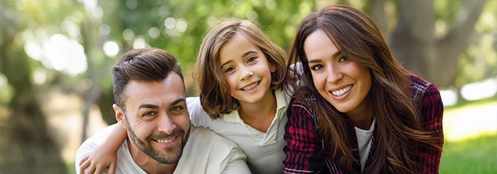 A family of three huddle together to smile for the camera with lush green trees behind them
