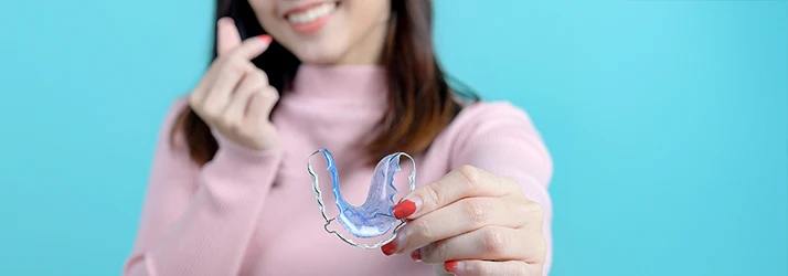 Woman in pink turtleneck with shoulder length brown hair gestures a heart symbol with her thumb and index finger while holding her retainer with the other hand Woman in pink turtleneck with shoulder length brown hair gestures a heart symbol with her thumb and index finger while holding her retainer with the other hand