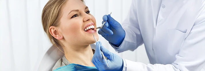 A blonde patient has her open mouth examined with a dental pick and mirror by two blue gloved hands A blonde patient has her open mouth examined with a dental pick and mirror by two blue gloved hands