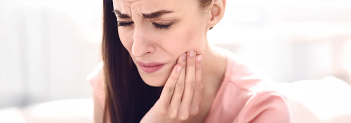 Woman with brown hair wearing a pink shirt scrunches her face in pain pressing her fingers to her cheek Woman with brown hair wearing a pink shirt scrunches her face in pain pressing her fingers to her cheek