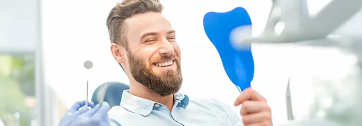 A blonde bearded man in a pale blue collared shirt gives a pleased crinkle eyed smile to his reflection in a hand held mirror A blonde bearded man in a pale blue collared shirt gives a pleased crinkle eyed smile to his reflection in a hand held mirror