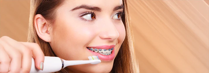 Close-up of a smiling young woman brushing her orthodontic braces with an electric toothbrush against a warm background. Close-up of a smiling young woman brushing her orthodontic braces with an electric toothbrush against a warm background.
