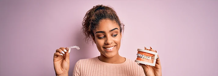 Smiling young woman holding clear aligners and a dental model with traditional braces, comparing orthodontic treatments. Smiling young woman holding clear aligners and a dental model with traditional braces, comparing orthodontic treatments.