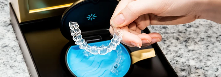 Close-up of a hand holding a clear Invisalign aligner above its storage case at an orthodontic office. Close-up of a hand holding a clear Invisalign aligner above its storage case at an orthodontic office.