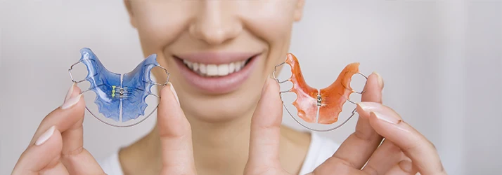Close-up of a smiling woman holding two colorful orthodontic retainers, showcasing dental care after braces. Close-up of a smiling woman holding two colorful orthodontic retainers, showcasing dental care after braces.