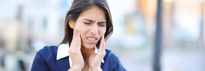 Woman experiencing jaw pain holding both sides of her face, indicating discomfort due to TMJ disorder. Woman experiencing jaw pain holding both sides of her face, indicating discomfort due to TMJ disorder.