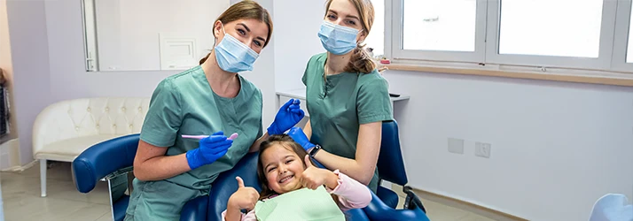 Happy child giving thumbs-up during preventative dental care visit with two pediatric dental professionals. Happy child giving thumbs-up during preventative dental care visit with two pediatric dental professionals.